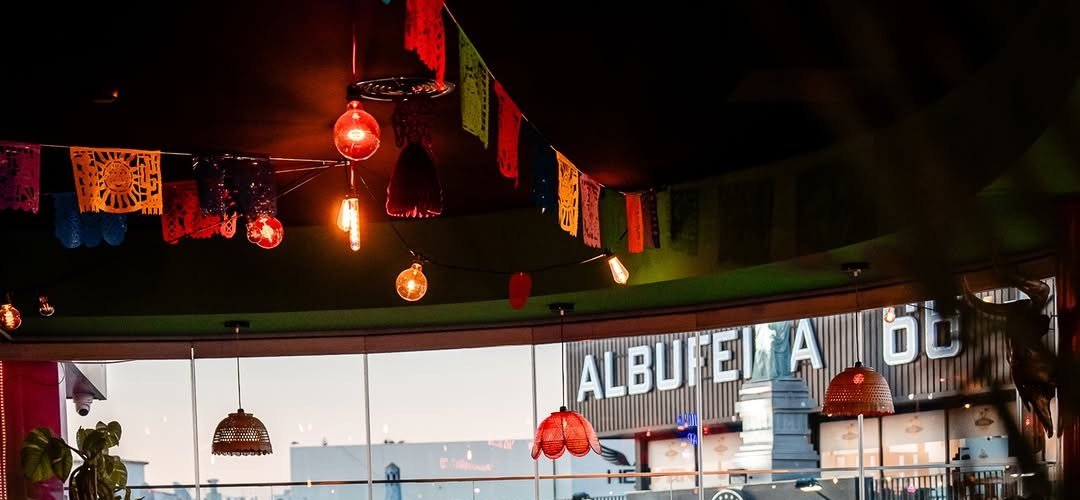 Sala com bandeirolas de papel recortado, lâmpadas acesas e janelas de vidro viradas para a avenida ao entardecer