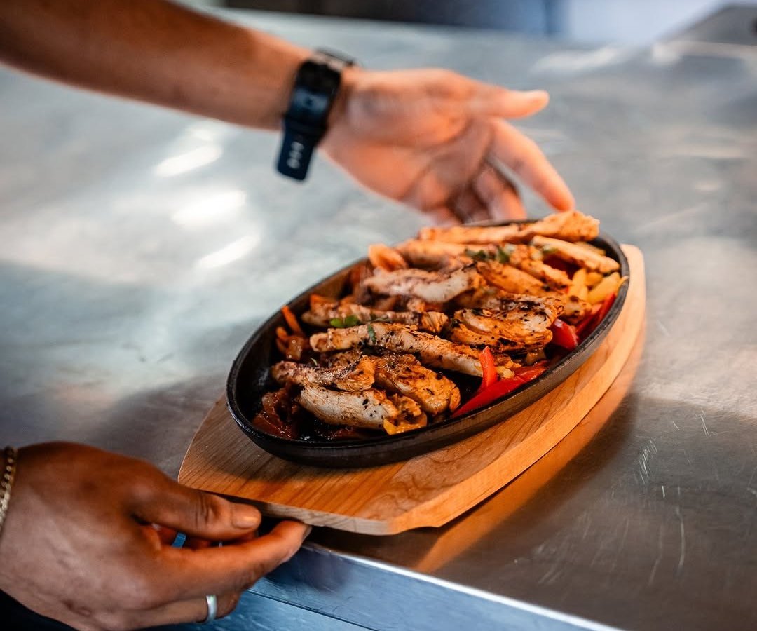 Fajitas de frango numa frigideira de ferro sobre tábua de madeira, seguradas por duas mãos